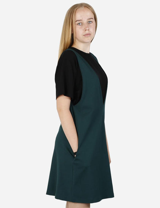 Woman in a pine green pinafore dress with a black t-shirt, side view, on a plain background.