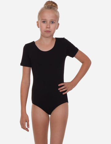 Child dancer in a black short sleeve leotard with a confident stance and hair in a bun, on a plain background.
